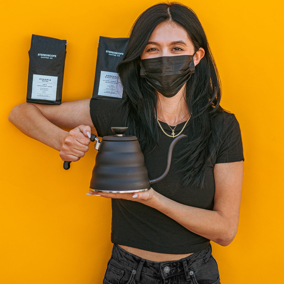 Model in all black with black mask and propping up two black bags of coffee on shoulder and elbow while holding a matte black metal gooseneck kettle and matching lid with plastic knob and black plastic covered handle against a yellow background.