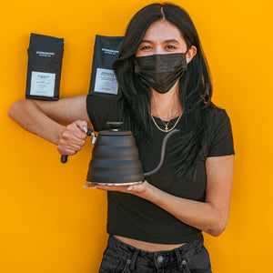 Model in all black with black mask and propping up two black bags of coffee on shoulder and elbow while holding a matte black metal gooseneck kettle and matching lid with plastic knob and black plastic covered handle against a yellow background.