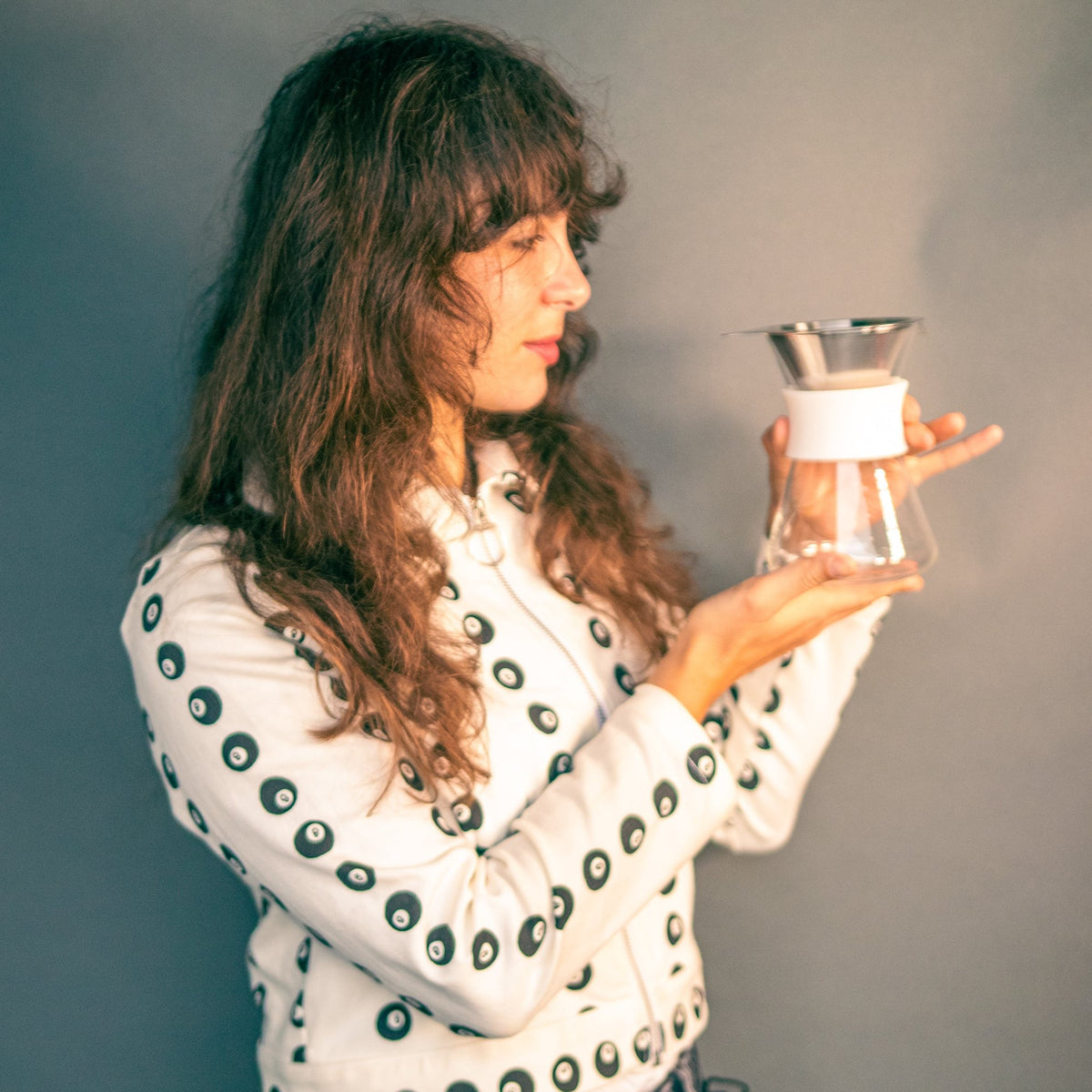 Woman holding a clear glass server with white silicone collar and stainless steel mesh filter against a grey background.