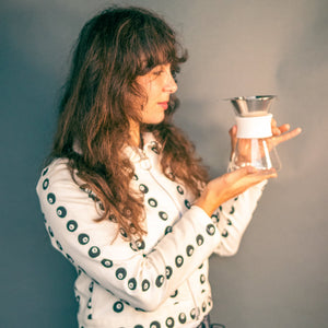 Woman holding a clear glass server with white silicone collar and stainless steel mesh filter against a grey background.