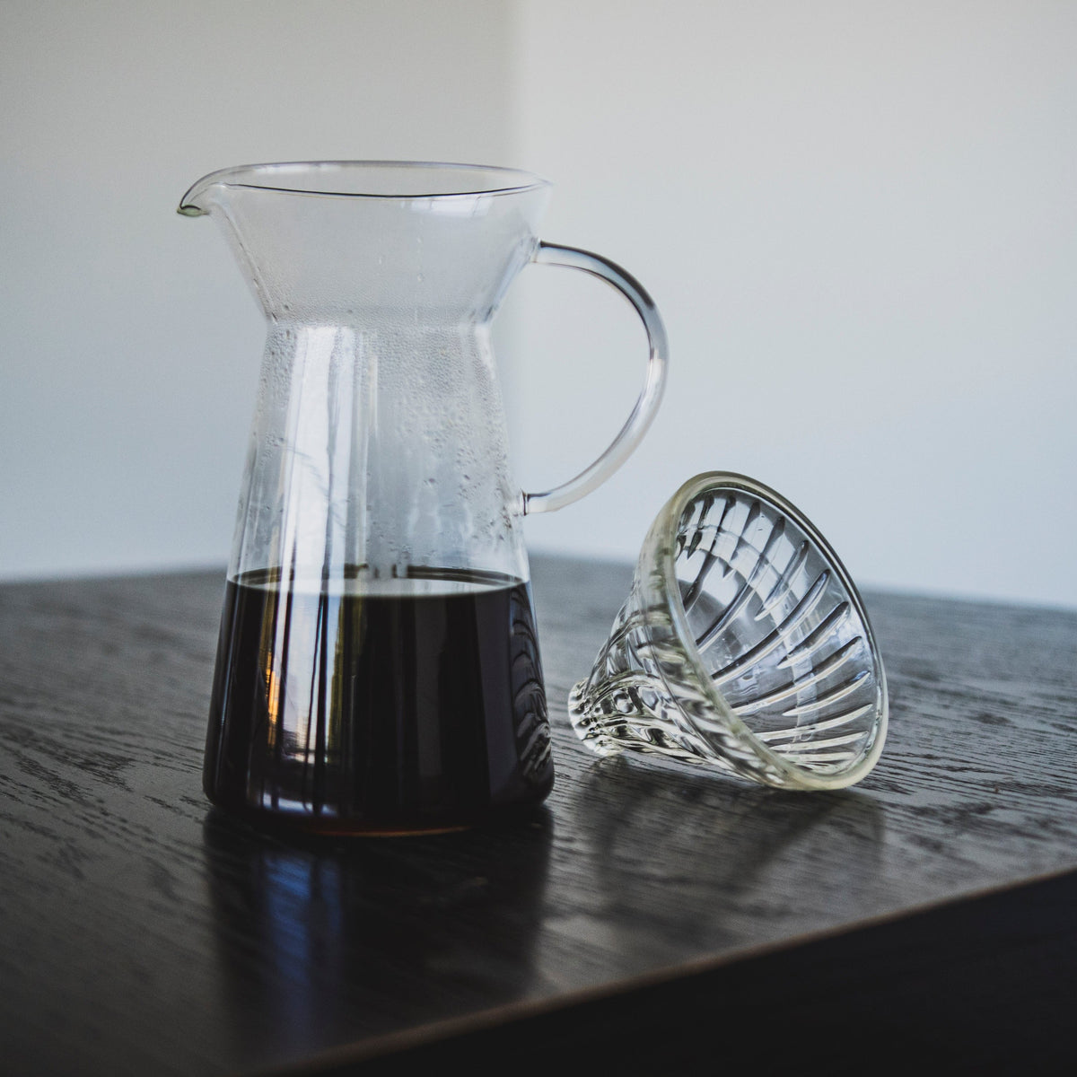 Tapered glass coffee server with fluted spout and all glass handle with coffee inside and a clear glass dripper with clear silicone ring seated beside on a dark wood table.