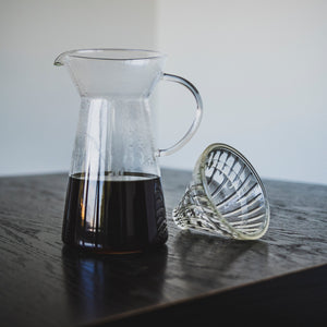 Tapered glass coffee server with fluted spout and all glass handle with coffee inside and a clear glass dripper with clear silicone ring seated beside on a dark wood table.