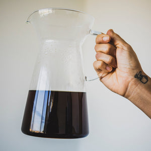 Model's hand holding a tapered clear glass server with fluted spout, all glass handle, and coffee inside.