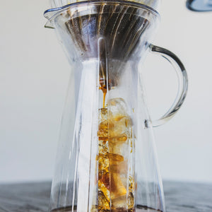 Coffee brewing over ice in a tall, tapered glass server with fluted spout and all glass handle with a white paper cone shaped filter in a glass cone shaped dripper with brewed coffee inside.
