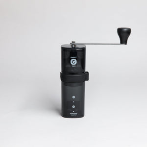 Black transparent plastic coffee mill with a slim metal handle attached to the shaft of the grinder.