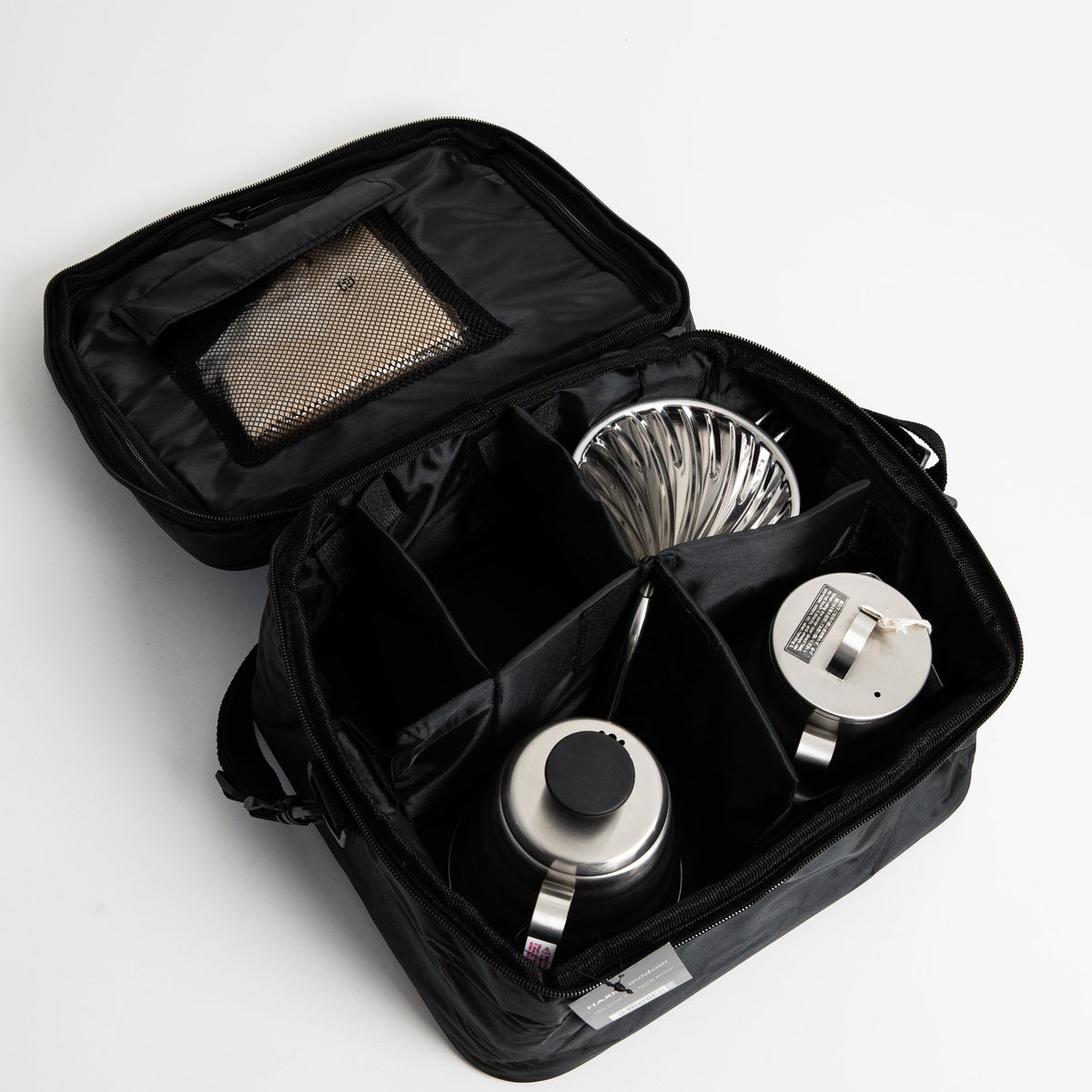 black carrier bag with compartments with stainless steel kettle, metal cone coffee dripper with a olive green base on top of a stainless steel coffee sever pot with stainless steel lid and coffee filters on a white background.