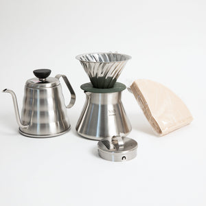stainless steel kettle, metal cone coffee dripper with a olive green base on top of a stainless steel coffee sever pot, stainless steel lid and coffee filters on a white background.