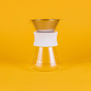 Clear glass server with white silicone collar and stainless steel mesh filter against a yellow background.
