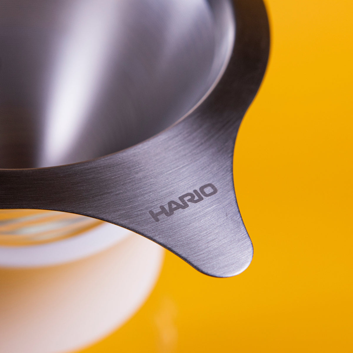 Stainless steel mesh filter with Hario logo on tab and blurred parts of the glass server with white silicone collar against a yellow background.