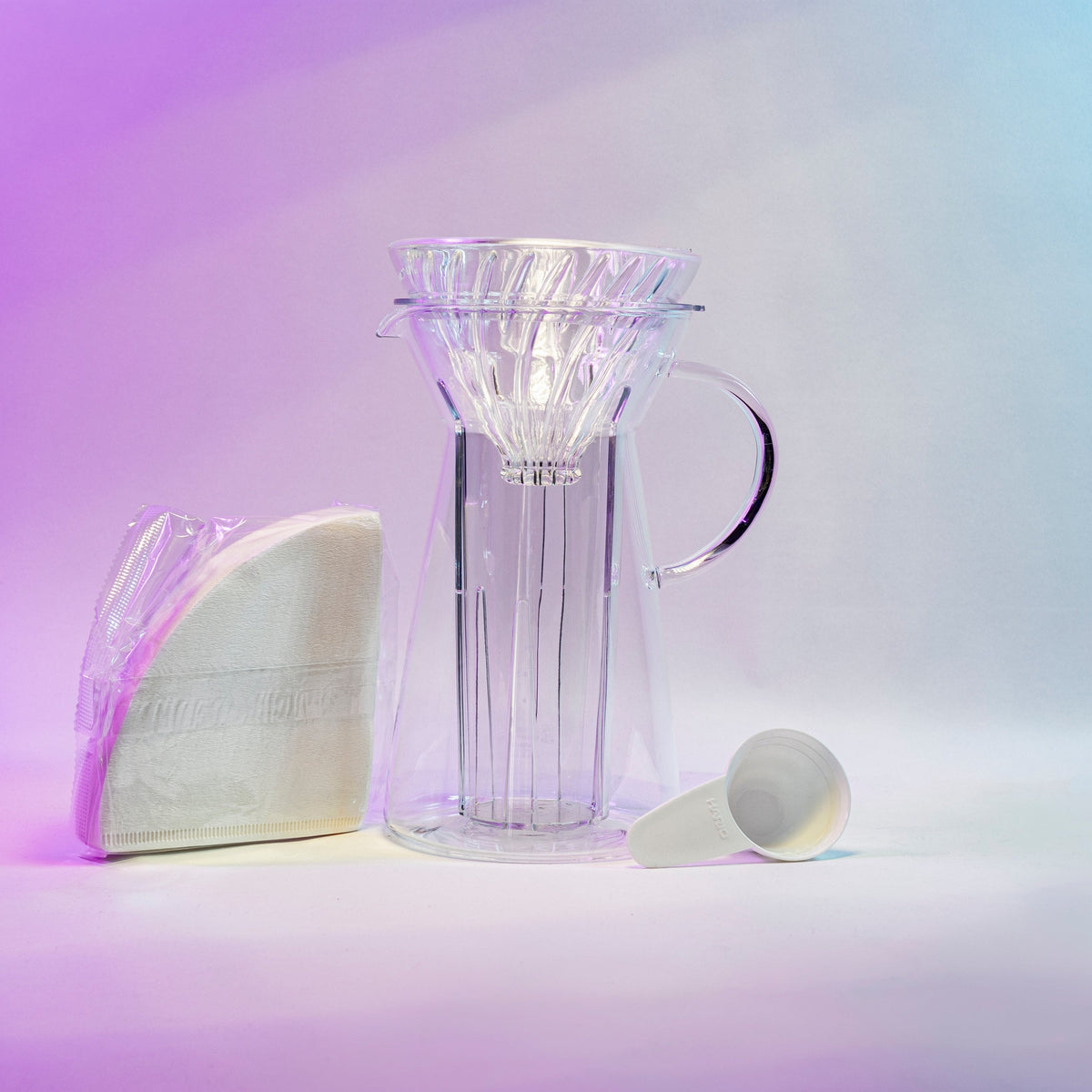 White cone shaped paper filters in a clear plastic bag next to a tapered glass dripper with fluted spout and all glass handle with a cone shaped glass dripper with ribs seated inside, and a white plastic coffee measuring spoon.
