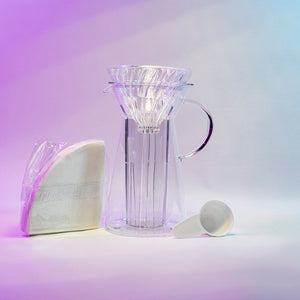 White cone shaped paper filters in a clear plastic bag next to a tapered glass dripper with fluted spout and all glass handle with a cone shaped glass dripper with ribs seated inside, and a white plastic coffee measuring spoon.