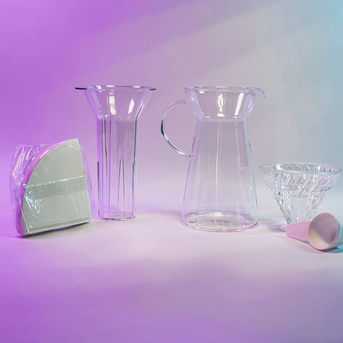 White cone shaped paper filters in a clear plastic bag next to a clear plastic ice insert, tapered glass server with fluted spout and all glass handle, glass cone shaped dripper with clear silicone collar, and white plastic measuring spoon.