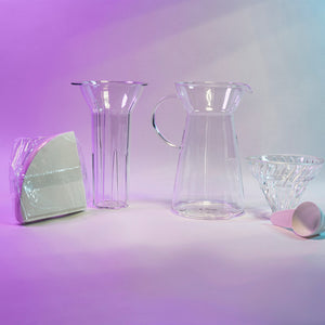 White cone shaped paper filters in a clear plastic bag next to a clear plastic ice insert, tapered glass server with fluted spout and all glass handle, glass cone shaped dripper with clear silicone collar, and white plastic measuring spoon.