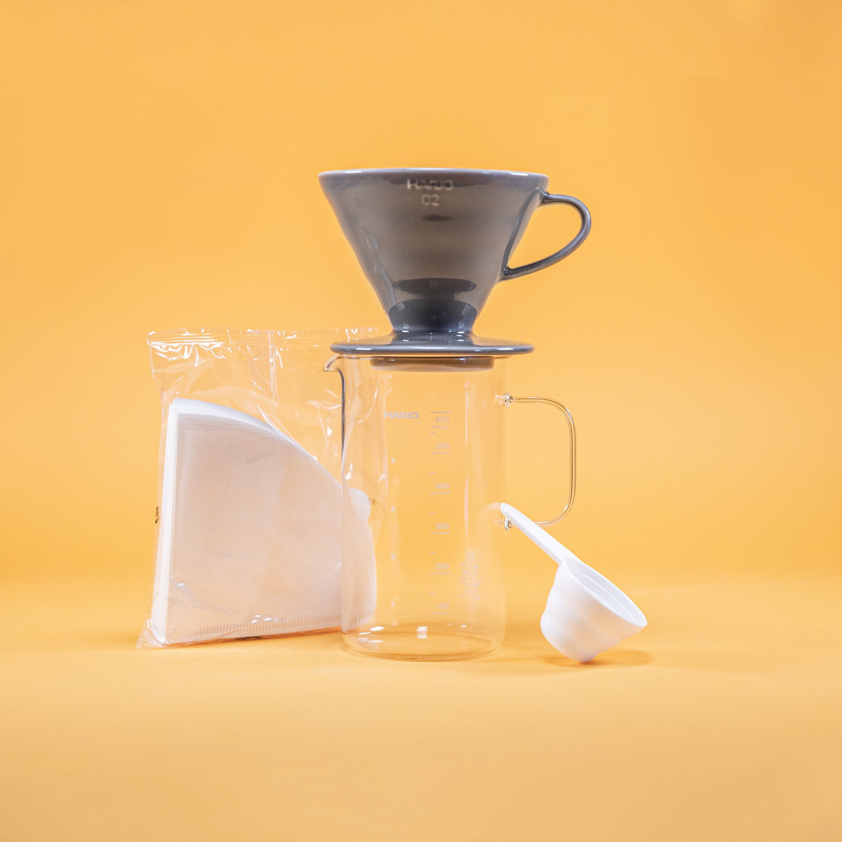Grey ceramic coffee dripper sitting atop glass coffee server with milliliter markings from 100 to 600 and full glass handle, white paper triangular shaped coffee filters in clear plastic bag, and white plastic coffee measuring spoon against a yellow background.