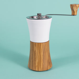 Hour glass shaped coffee hand grinder with olive wood base, white ceramic neck, stainless steel grinder assembly and screw, stainless steel handle with olive wood knob, and set against a blue background.