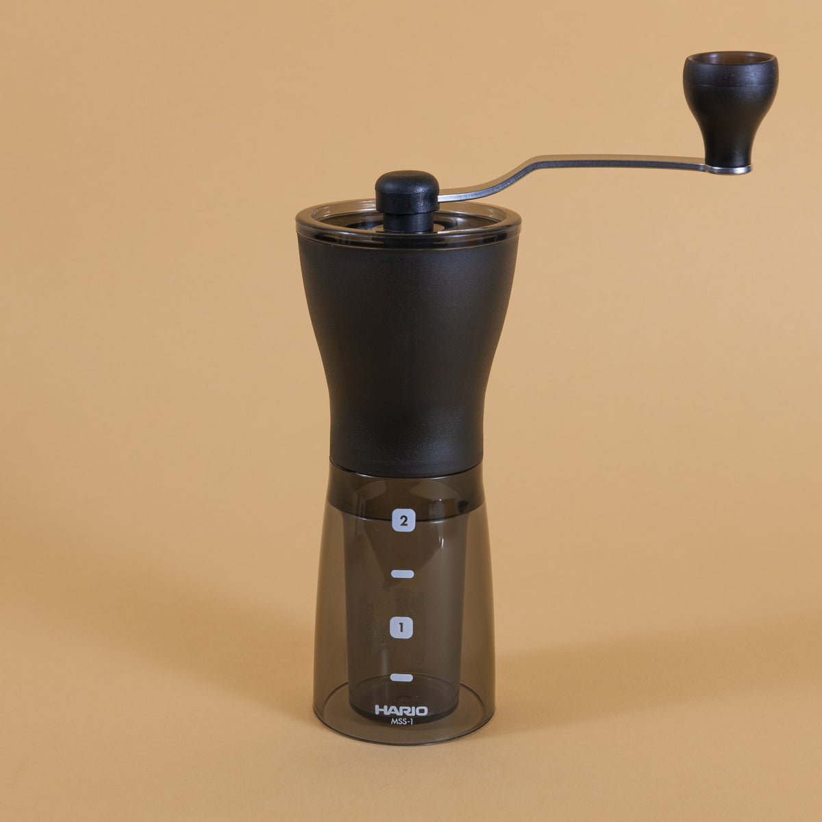 Slim black plastic Coffee Mill with handle atop a dark transparent plastic container with two level markings on an orange backdrop