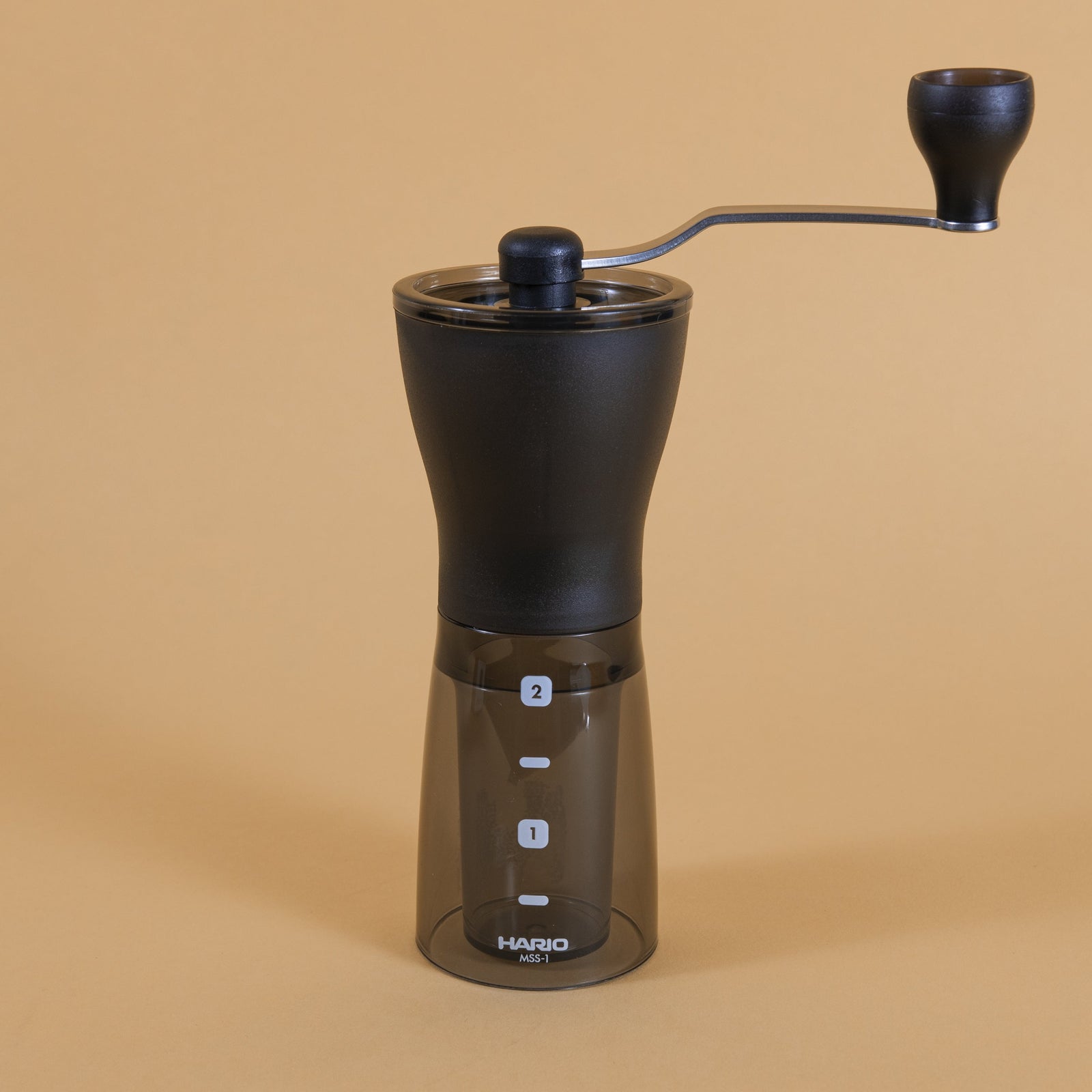 Slim black plastic Coffee Mill with handle atop a dark transparent plastic container with two level markings on an orange backdrop