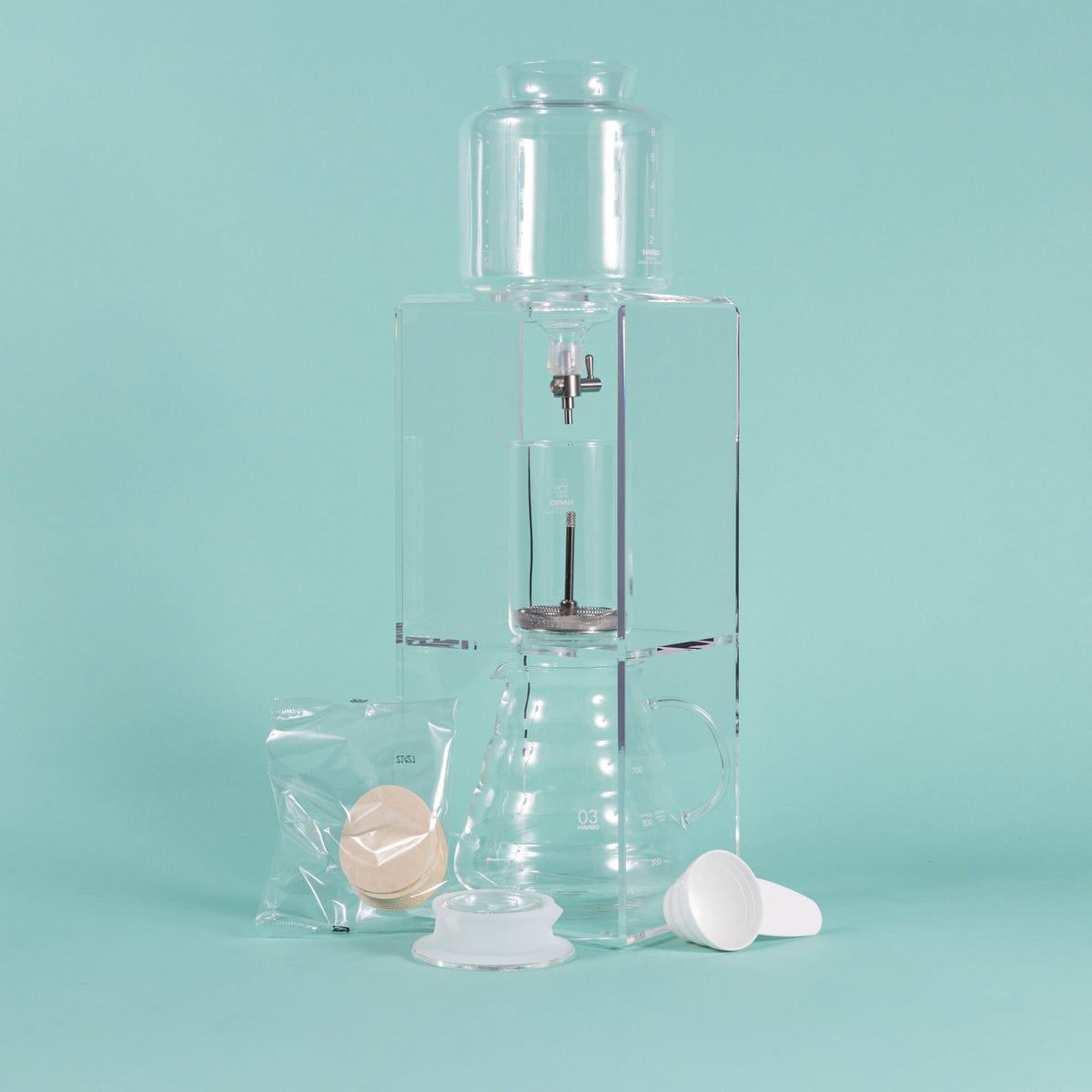 Tall clear acrylic tower stand with water reservoir and drip spout sitting over a glass cylinder with metal mesh filter for coffee grounds, with all glass coffee server with handle underneath and paper filter pack, server lid, and white plastic scoop on a teal background